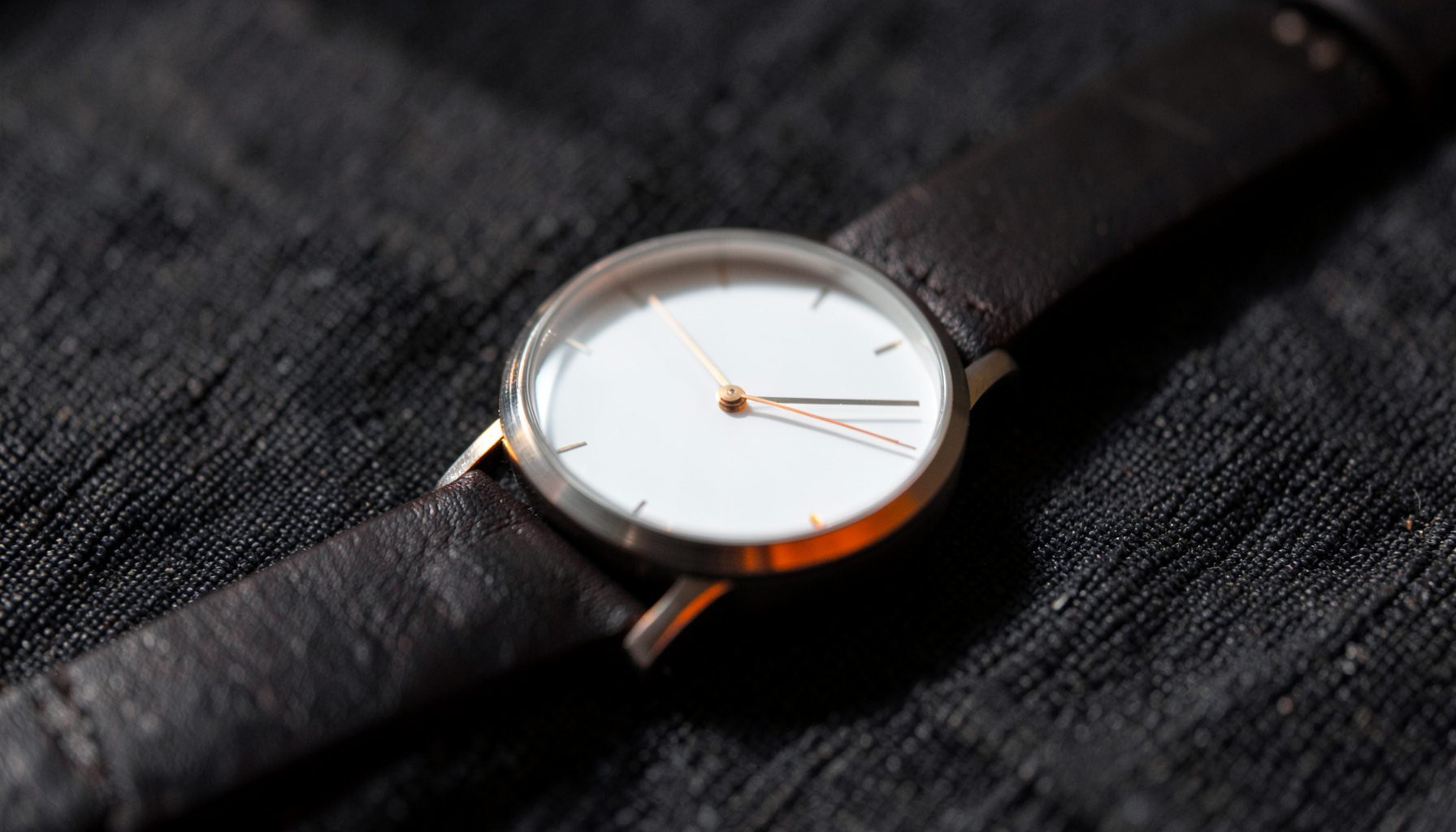 Close-up of a minimalist wristwatch with a dark leather strap and a clean white dial, resting on a textured charcoal fabric surface in soft directional light.